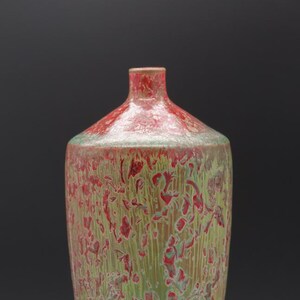 May include: A ceramic vase with a narrow neck and a rounded body, featuring a mottled glaze pattern of vibrant red and light green colors. The surface texture appears layered with vertical streaks and irregular patches of color throughout the piece.