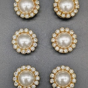 May include: Six round, decorative buttons with a central pearl surrounded by a ring of small, clear crystals. The crystals are set in a gold-colored metal frame. The buttons are arranged on a gray surface.
