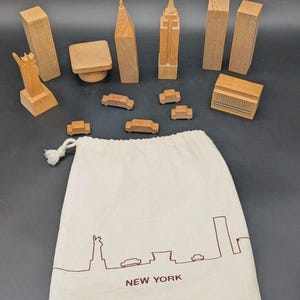 May include: A set of wooden New York City building and vehicle blocks, including the Statue of Liberty, skyscrapers, and cars, accompanied by a cream-coloured fabric drawstring storage bag featuring a brown line-art skyline and the text NEW YORK.