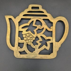 May include: A gold-colored metal trivet in the shape of a teapot. The trivet features a decorative cutout design of leaves and grapes. The handle and spout are clearly defined, and the lid has a rectangular opening.