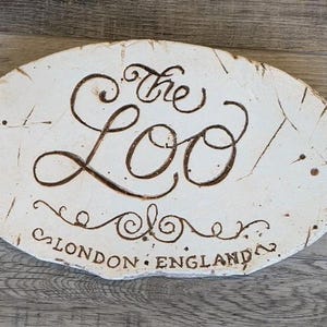 May include: Oval, off-white decorative sign with the words "The Loo" in a brown, cursive font. The sign also includes the text "LONDON ENGLAND" in a smaller, brown font. The sign has a distressed, rustic appearance.