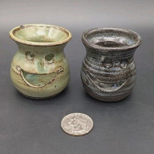 May include: Two small, handmade ceramic vases with smiling faces. One vase is light green with brown accents, the other is dark gray with blue and brown streaks. A U.S. quarter is next to the vases for scale.