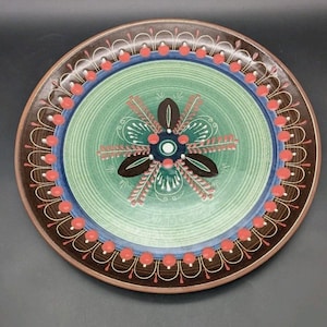 May include: A round ceramic plate featuring a central dark brown floral motif on a light green background. The design includes a blue circular border accented with a ring of small red dots and white line arches against a dark brown rim.