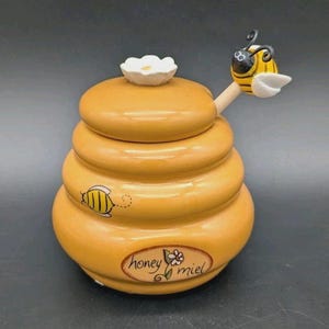 May include: A yellow ceramic honey pot shaped like a beehive with a lid featuring a white flower. A wooden dipper with a bee topper rests inside. The pot has a painted bee and an oval label with the text honey miel and a small flower illustration.