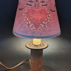 May include: A table lamp featuring a dark brown wooden spool base and a conical lampshade in a deep maroon color. The lampshade is decorated with a cut-out pattern of a bow, a heart, and leafy branches, allowing light to shine through the design.