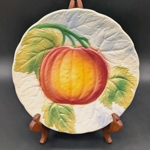 May include: A round ceramic plate featuring a raised, three-dimensional relief of a central orange and yellow pumpkin surrounded by green and white embossed leaves. The plate has a scalloped edge and is displayed on a wooden stand.
