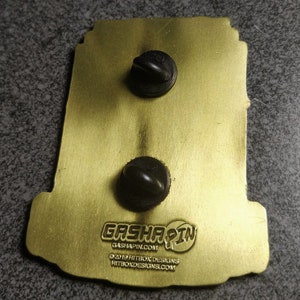 May include: Gold-colored metal pin with a black and white illustration of a robot. The pin has two black metal clasps on the back and the text "Gashapin" with the website "gashapin.com". The text "&copy;2019 Hitbox Designs" and the website "hitboxdesigns.com" are also on the back of the pin.