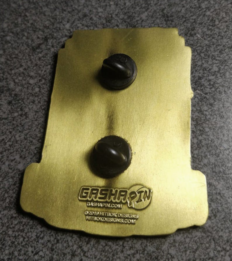 May include: Gold-colored metal pin with a black and white illustration of a robot. The pin has two black metal clasps on the back and the text "Gashapin" with the website "gashapin.com". The text "&copy;2019 Hitbox Designs" and the website "hitboxdesigns.com" are also on the back of the pin.
