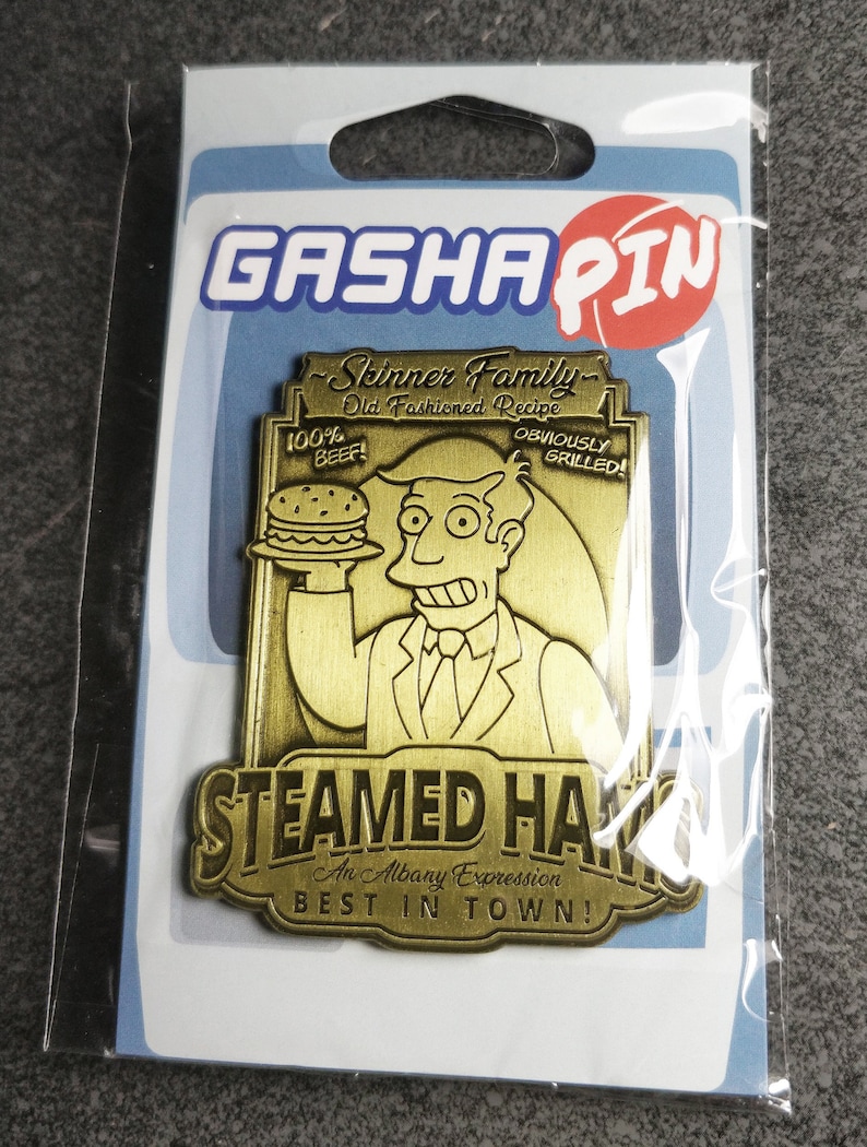 May include: A gold-colored metal pin featuring a cartoon character holding a hamburger. The pin reads "Skinner Family - Old Fashioned Recipe - 100% Beef - Obviously Grilled!" and "Steamed Hams - An Albany Expression - Best in Town!"