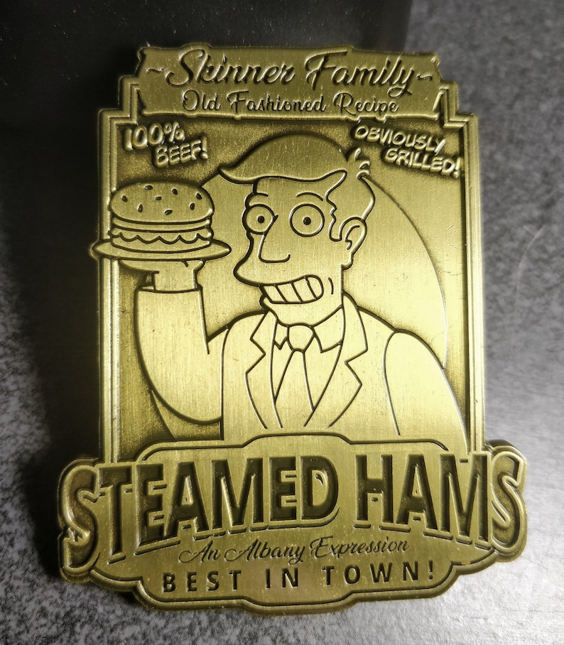 May include: A gold-colored metal pin featuring a cartoon character holding a hamburger. The pin reads "Skinner Family Old Fashioned Recipe 100% BEEF! OBVIOUSLY GRILLED!" and "STEAMED HAMS An Albany Expression BEST IN TOWN!"