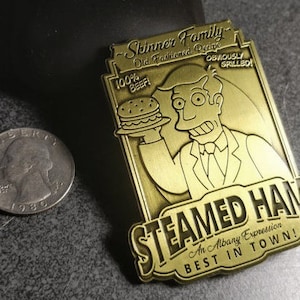 May include: A gold-colored metal pin featuring a cartoon character holding a hamburger. The pin reads "Skinner Family Old Fashioned Recipe 100% Beef! Obviously Grilled! Steamed Hams An Albany Expression Best in Town!"