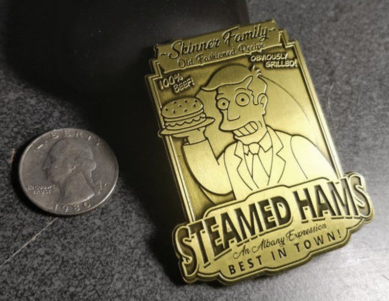 May include: A gold-colored metal pin featuring a cartoon character holding a hamburger. The pin reads "Skinner Family Old Fashioned Recipe 100% Beef! Obviously Grilled! Steamed Hams An Albany Expression Best in Town!"