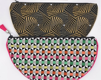 May include: Two half-moon shaped zippered pouches with different geometric patterns. The top pouch has a black and gold geometric pattern. The bottom pouch has a black, white, green, pink, and orange geometric pattern. Both pouches have a pink zipper and a black tassel.