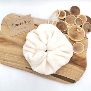 Puede incluir: Un scrunchie redondo y esponjoso de color blanco con un lazo en la parte superior. El scrunchie est&aacute; sobre una superficie de madera con una tarjeta de visita que dice "Cousettes - Editions couture artisanales - www.cousettesjewelry.com".