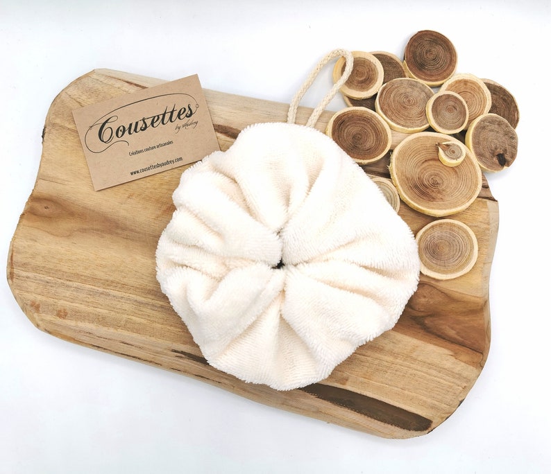 Puede incluir: Un scrunchie redondo y esponjoso de color blanco con un lazo en la parte superior. El scrunchie est&aacute; sobre una superficie de madera con una tarjeta de visita que dice "Cousettes - Editions couture artisanales - www.cousettesjewelry.com".