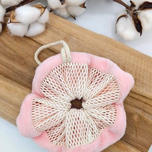 Puede incluir: Un puf de ba&ntilde;o de malla rosa y blanco con cierre de cord&oacute;n. El puf est&aacute; sobre una superficie de madera.