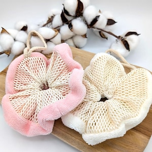 Puede incluir: Dos esponjas de ba&ntilde;o, una rosa y otra blanca, con textura de malla y una capa exterior suave y esponjosa. Las esponjas est&aacute;n sobre una superficie de madera.