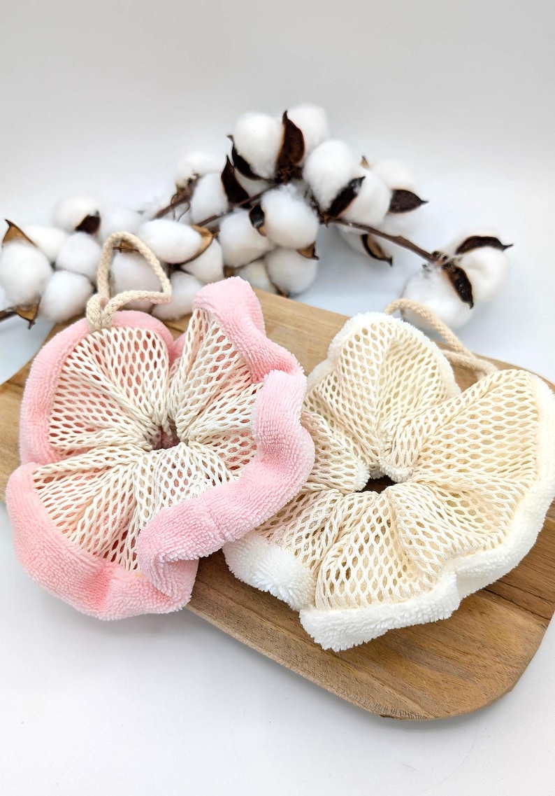 Puede incluir: Dos esponjas de ba&ntilde;o, una rosa y otra blanca, con textura de malla y una capa exterior suave y esponjosa. Las esponjas est&aacute;n sobre una superficie de madera.