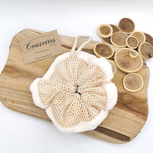 Puede incluir: Un puff de ba&ntilde;o blanco y beige con textura de malla y cierre de cord&oacute;n. El puff est&aacute; sobre una superficie de madera con discos de madera.