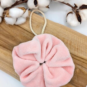 Puede incluir: Un puff de ba&ntilde;o rosa en forma de flor con un lazo de cuerda blanco. El puff est&aacute; sobre una superficie de madera.