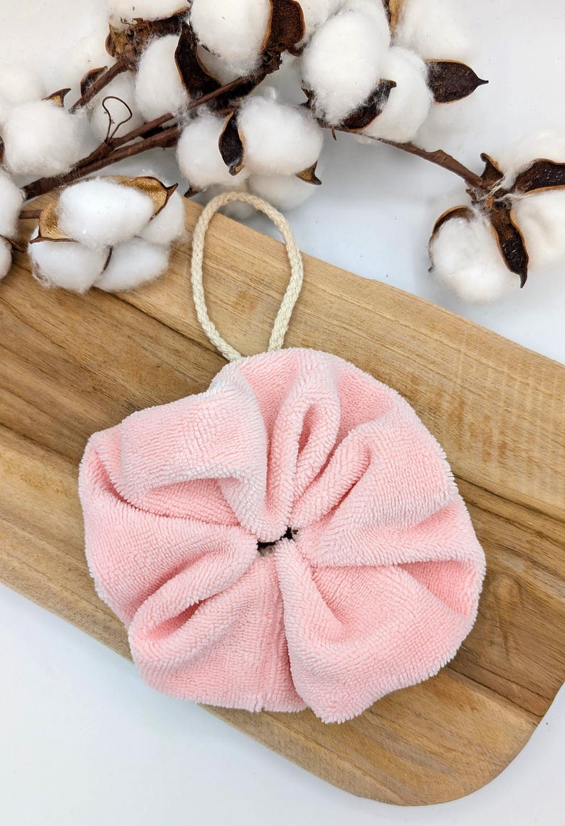 Puede incluir: Un puff de ba&ntilde;o rosa en forma de flor con un lazo de cuerda blanco. El puff est&aacute; sobre una superficie de madera.