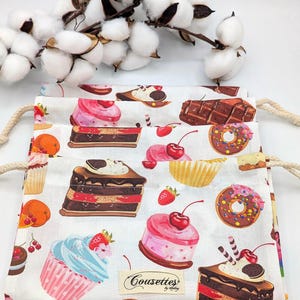 May include: A white drawstring bag with a colorful pattern of cupcakes, donuts, and cakes. The bag has a label that says "Cousettes by Melody".