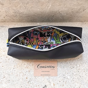 Può includere: Borsa nera con cerniera con una cerniera bianca e una fodera con stampa grafica colorata. La fodera presenta una varietà di parole e immagini colorate, tra cui "Best", "Ok" e un segno di pace. La borsa è aperta e la fodera è visibile. La borsa è etichettata "Cousettes" su un piccolo cartoncino nell'angolo in basso a destra.