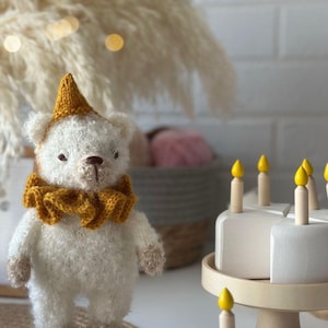 May include: A handmade white teddy bear wearing a yellow knitted hat and collar. The bear stands near a wooden cake stand with white cake slices topped with yellow candle-shaped decorations. A decorative item for a nursery or child's room.