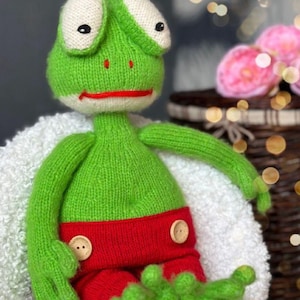 May include: A handmade green knitted frog doll with white eyes, a red mouth, and red shorts. The frog has button details and is sitting on a white surface. The doll is a whimsical, handcrafted item.
