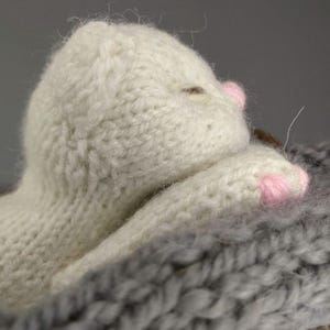 Op de afbeelding: Een handgemaakt, crèmekleurig gebreid kattenspeeltje met roze accenten op de poten. De kat ligt in een grijze gebreide deken. Het speelgoed is gemaakt van zacht garen.