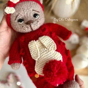 May include: A handmade knitted teddy bear wearing a red outfit with a white bunny applique. The bear has a brown face, a red hood with white bows, and is held in someone's hands. The text "By Ola Osipova" is visible.