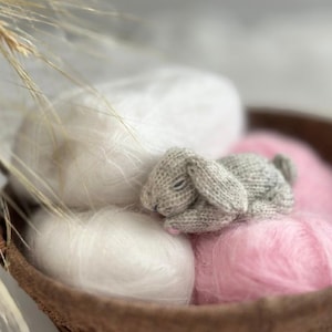 May include: A small, gray knitted bunny rests on a bed of pink and white yarn inside a brown woven basket. The bunny has its eyes closed, and the yarn appears soft and fluffy. Dried grasses are in the background.