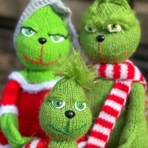 May include: Three knitted Grinch dolls. The dolls are green with black noses and green eyes. One doll wears a red and white striped scarf, another wears a red and white striped shirt, and the third wears a gray hat.