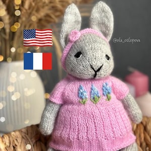 Könnte beinhalten: Ein graues gestricktes Häschen, das ein rosa Kleid mit blauen gestickten Blumen trägt. Das Häschen sitzt in einem geflochtenen Korb. Das Bild enthält auch den Text "@ola_oslopova".