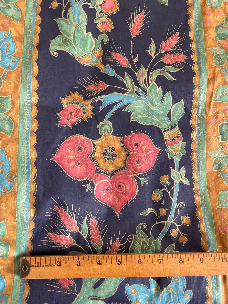 May include: A navy blue fabric with a gold, pink, and green floral pattern. The fabric has a border with a gold, brown, and green pattern. A ruler is placed on the fabric to show the scale.