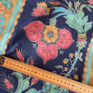 May include: A close-up of a dark blue fabric with a gold, teal, and red floral pattern. A wooden ruler is placed on top of the fabric, with the numbers 2 through 20 visible.