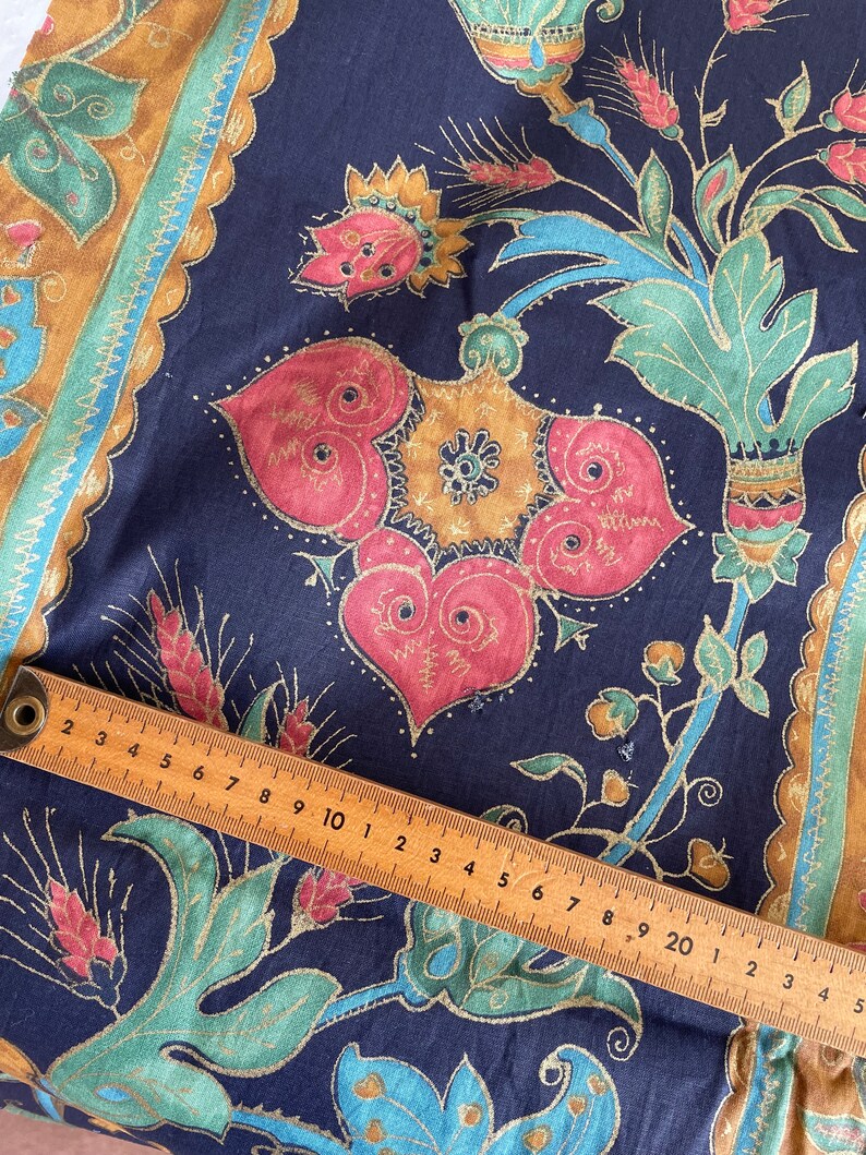 May include: A close-up of a dark blue fabric with a gold, teal, and red floral pattern. A wooden ruler is placed on top of the fabric, with the numbers 2 through 20 visible.