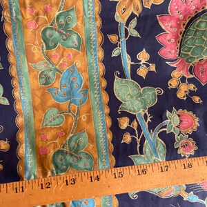 May include: A close-up of a fabric with a repeating pattern of gold, green, and blue floral designs on a dark blue background. A wooden ruler with inch markings is placed on top of the fabric.