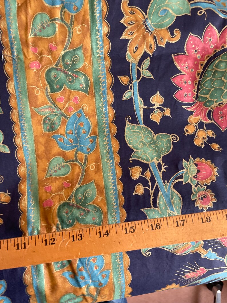 May include: A close-up of a fabric with a repeating pattern of gold, green, and blue floral designs on a dark blue background. A wooden ruler with inch markings is placed on top of the fabric.
