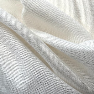 Puede incluir: Primer plano de una tela suave de color blanco roto con un tejido texturizado. La tela está drapeada en pliegues suaves, creando sombras y luces. El patrón de tejido es visible, agregando profundidad y detalle al textil.