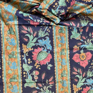 May include: A close-up of a dark blue fabric with a repeating pattern of stylized flowers and wheat stalks. The fabric has a gold and green striped border.