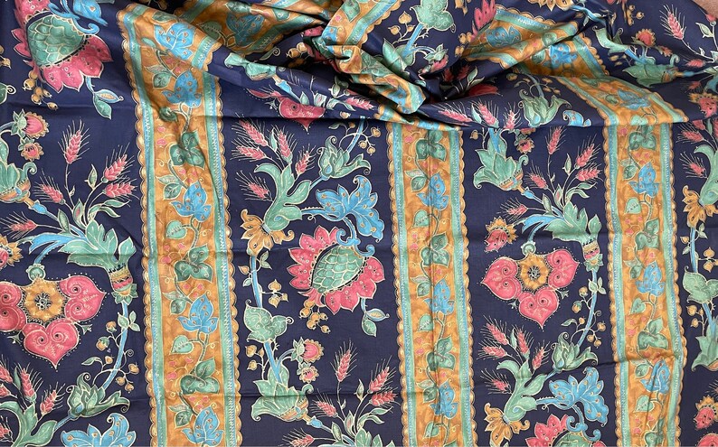 May include: A close-up of a dark blue fabric with a repeating pattern of stylized flowers and wheat stalks. The fabric has a gold and green striped border.