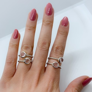 Puede incluir: Dos anillos de plata con una piedra preciosa redonda y transparente en cada uno. Los anillos se llevan en el índice y el dedo medio de una mano con esmalte de uñas rosa.