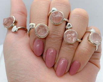 May include: Five silver rings with pink crystal stones set in a heart shape. The rings are worn on a hand with pink nail polish.