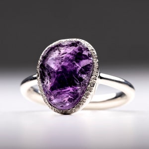 Könnte beinhalten: Ein silberner Ring mit einem großen, facettierten, violetten Amethyst-Edelstein, der in einer Zargenfassung mit einer Reihe kleiner Diamanten um den Rand herum gefasst ist.