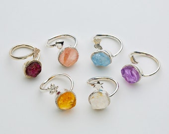 May include: Six silver rings with different coloured gemstones. Each ring has a small decorative charm attached to the band. The gemstones are pink, blue, purple, yellow, and white.