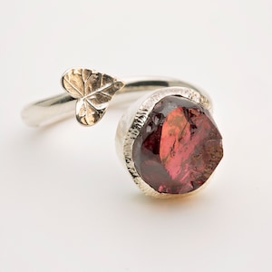 May include: A silver ring with a red gemstone and a gold leaf design. The ring is open and can be adjusted to fit different finger sizes.