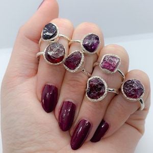 May include: A set of eight silver rings with raw, natural, pink and purple gemstones. The rings are worn on a hand with dark red nail polish.