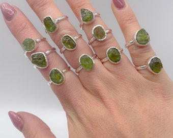 May include: A collection of 12 silver rings with raw green peridot stones. The rings are displayed on a hand wearing pink nail polish.