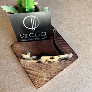 May include: A hair clip with a tortoiseshell pattern, black accents, and a silver clasp rests on a wooden square. A black card with the logo "Loctia STAY AND PROTECT" is propped up behind it. A small potted plant is in the background.
